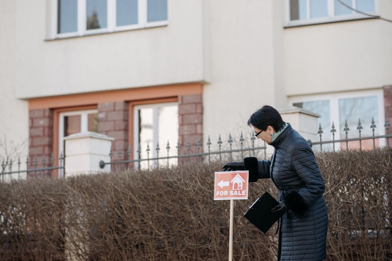 testimonials-img Real estate agent in a black coat placing a 'For Sale' sign in front of a house.